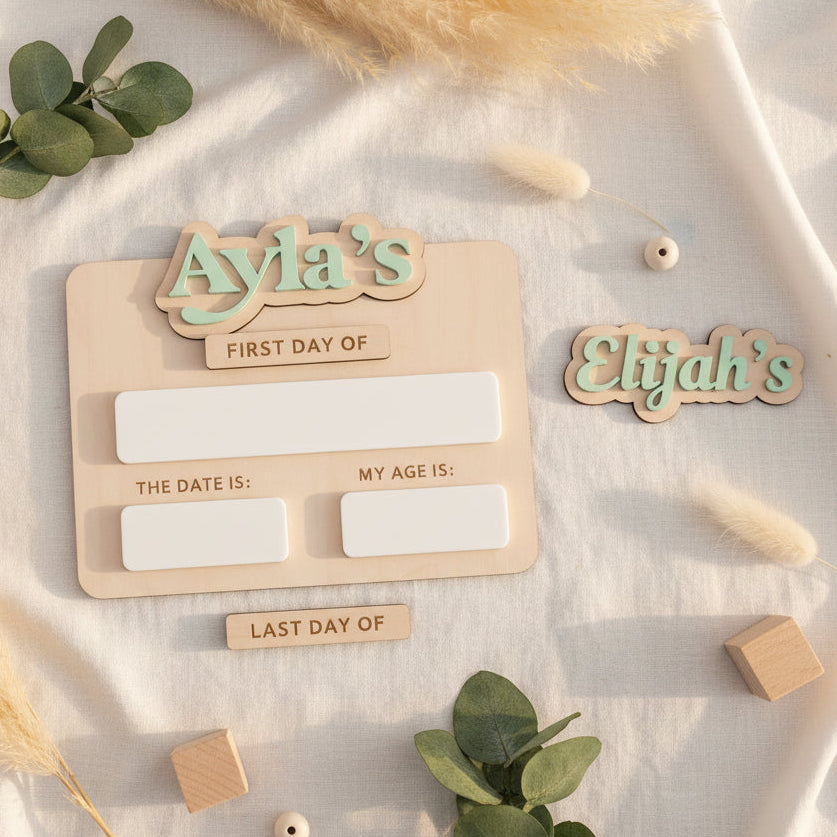 Wooden milestone board with 'Ayla's First Day of' and 'Elijah's' text, surrounded by decorative elements on a light fabric background.