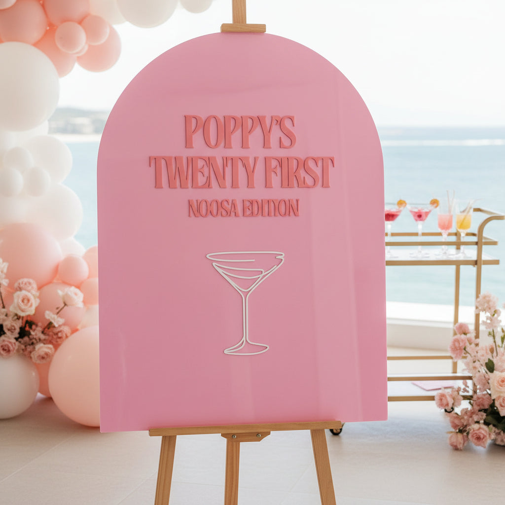 Pink event sign with 'Poppy's Twenty First Noosa Edition' text and martini glass design on a white surface.
