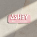 Pink name tag with 'ASHBY ENROLLED NURSE' text on a light gray background