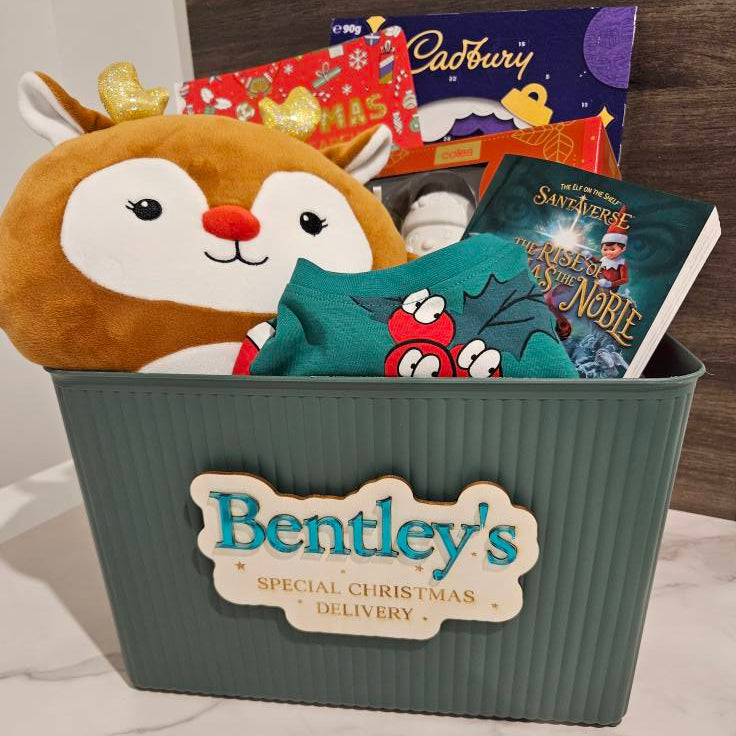 Gift basket with plush deer, books, and Cadbury chocolate labeled 'Bentley's Special Christmas Delivery' on a wooden surface.