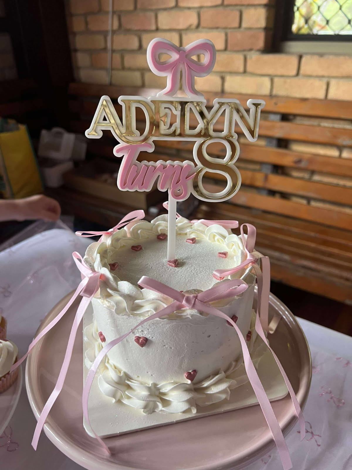 White cake with pink ribbons and a 'Adeyn Turning 8' topper on a table.