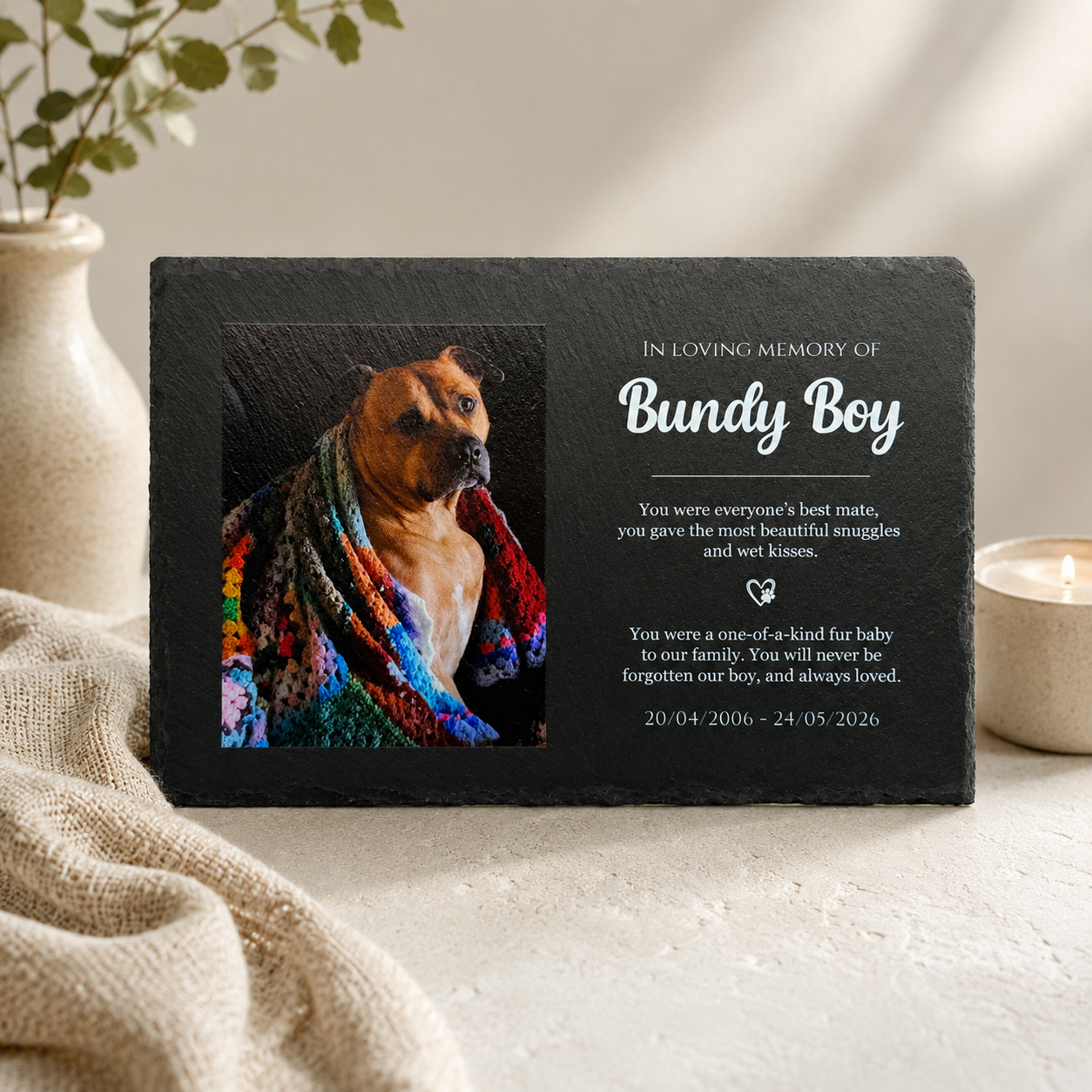 Memorial plaque for a dog named Bundy Boy with a photo of the dog wrapped in a colorful blanket, placed on a surface with a candle and plant in the background.