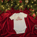 Baby onesie with 'My first Christmas' text on a textured surface with Christmas decorations.