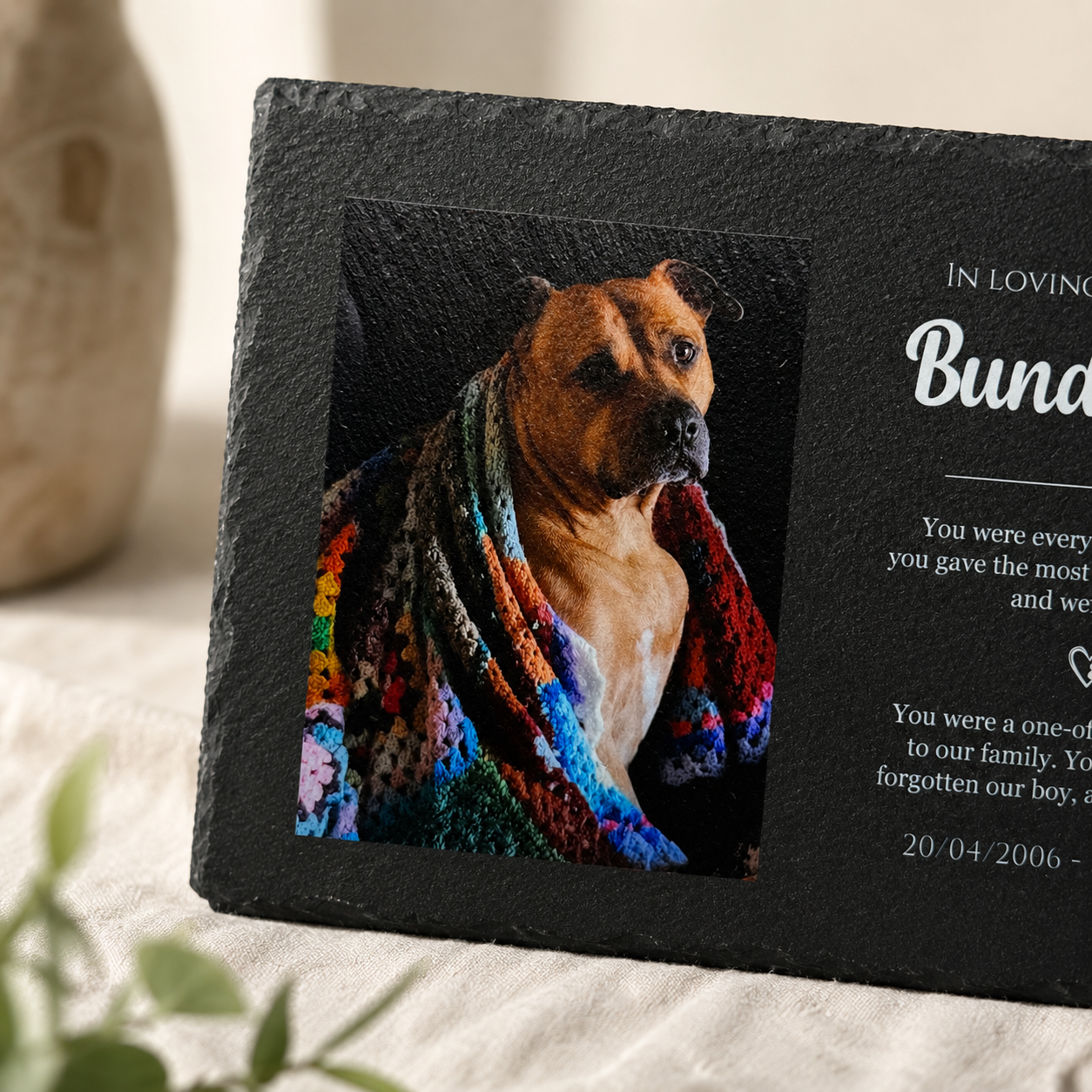 Memorial plaque with a dog wrapped in a colorful blanket and personalized text on a soft surface.