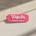 Pink name tag with 'Ursula Families Pastor' text on a wooden background