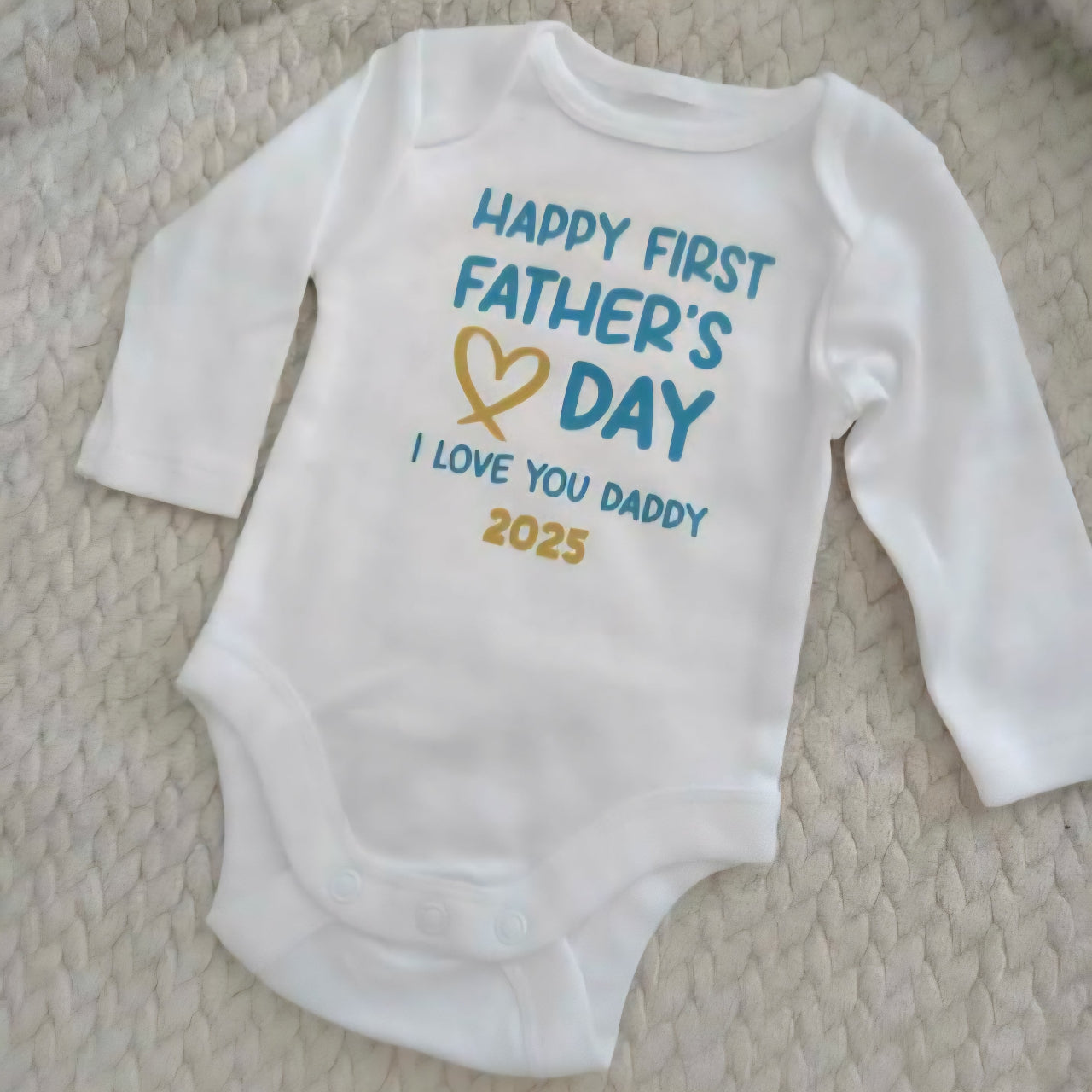 White baby onesie with 'Happy First Father's Day I Love You Daddy 2025' text on a textured surface