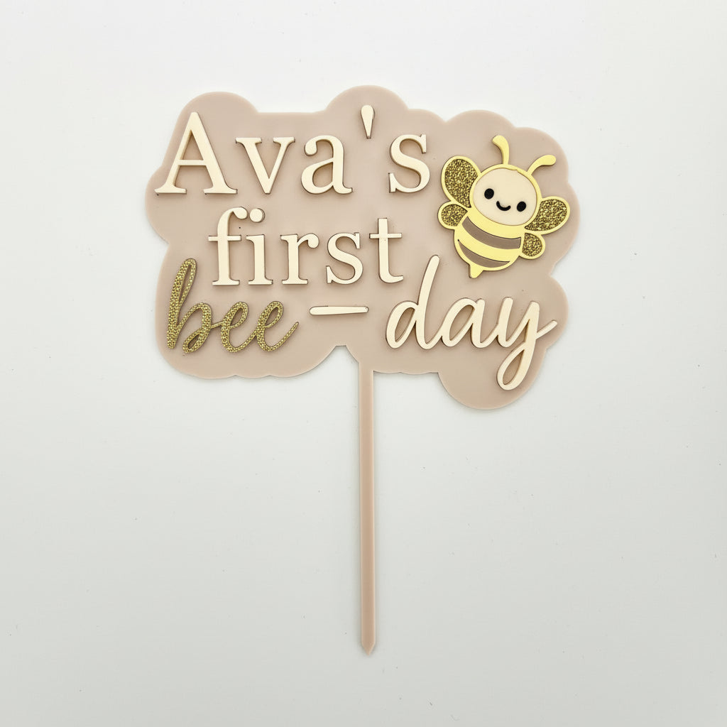 Decorative cake topper with 'Ava's first bee-day' text and a bee illustration on a white background.