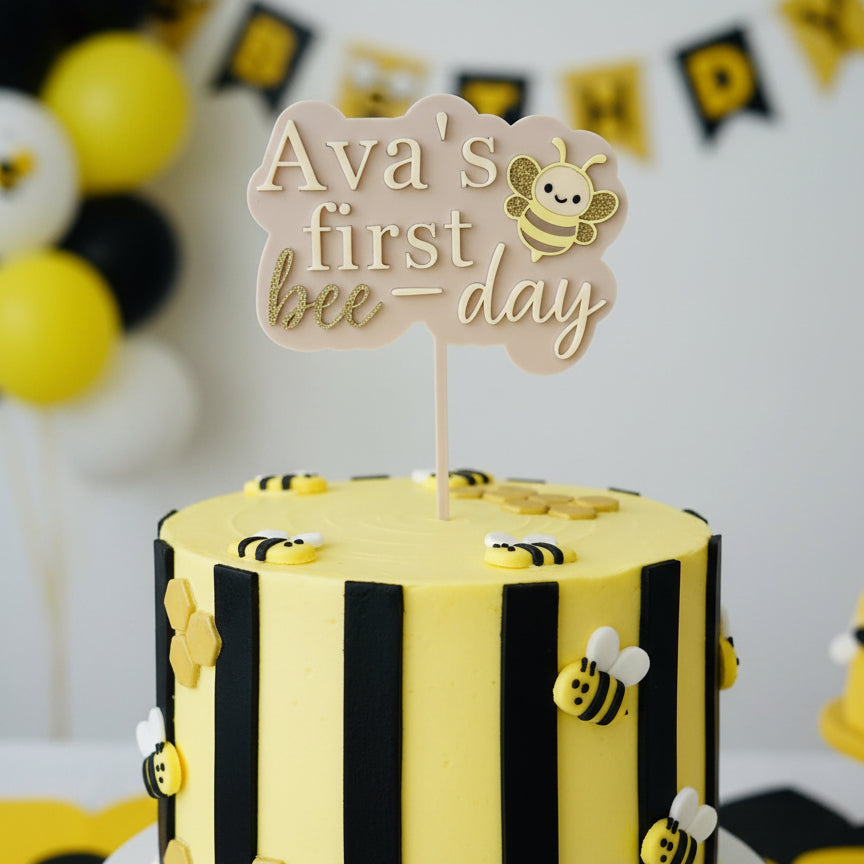 Decorative cake topper with 'Ava's first bee-day' text and a bee illustration on a white background.