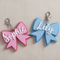 Two bow-shaped keychains, one pink and one blue, with 'Sophie' and 'Lily' engraved on them, on a textured fabric background.