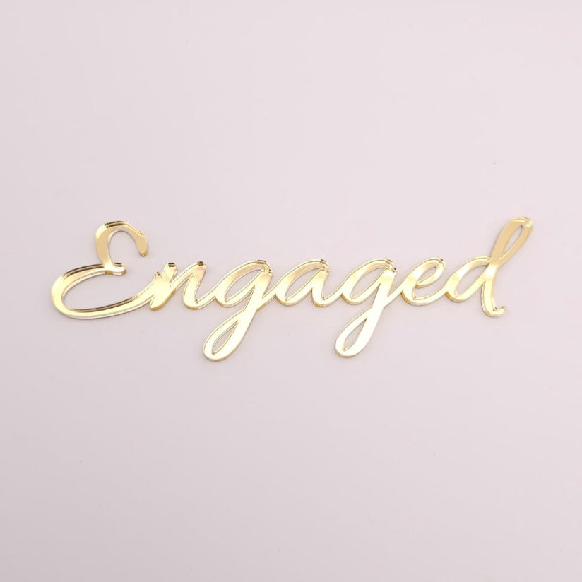 Gold 'Engaged' flat lay cake topper on a light pink background