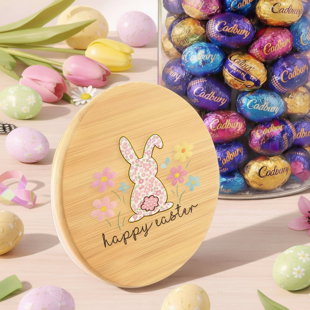 Glass jar with wooden lid filled with Cadbury eggs, surrounded by Easter decorations on a light background.