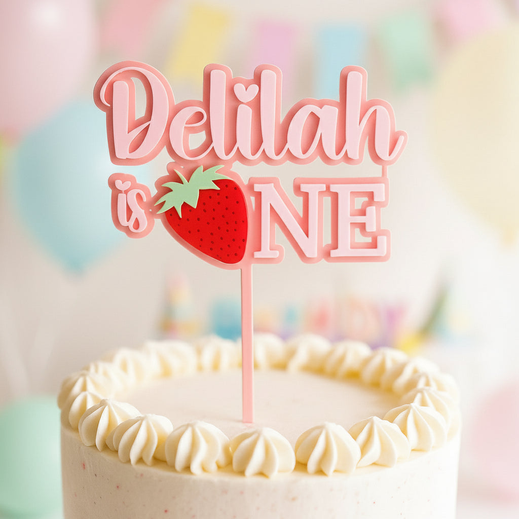Cake topper with 'Delilah is ONE' and a strawberry on a cake.